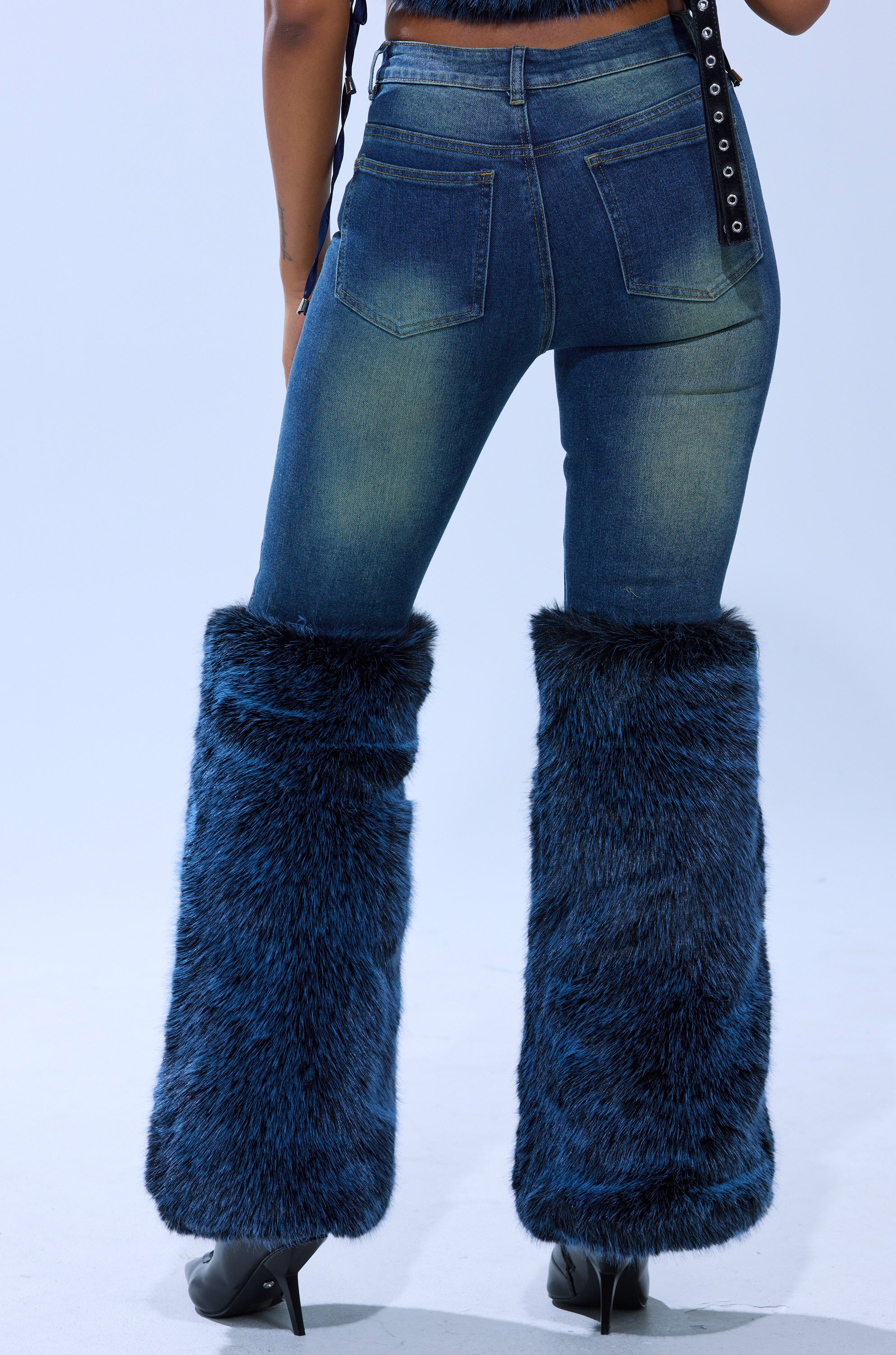 A person wears the ALL A DREAM DENIM PANT WITH FUR CUFFS, featuring high-rise jeans and faux fur cuffs at the lower legs, paired with black heels. The photo is taken from behind against a plain light background.