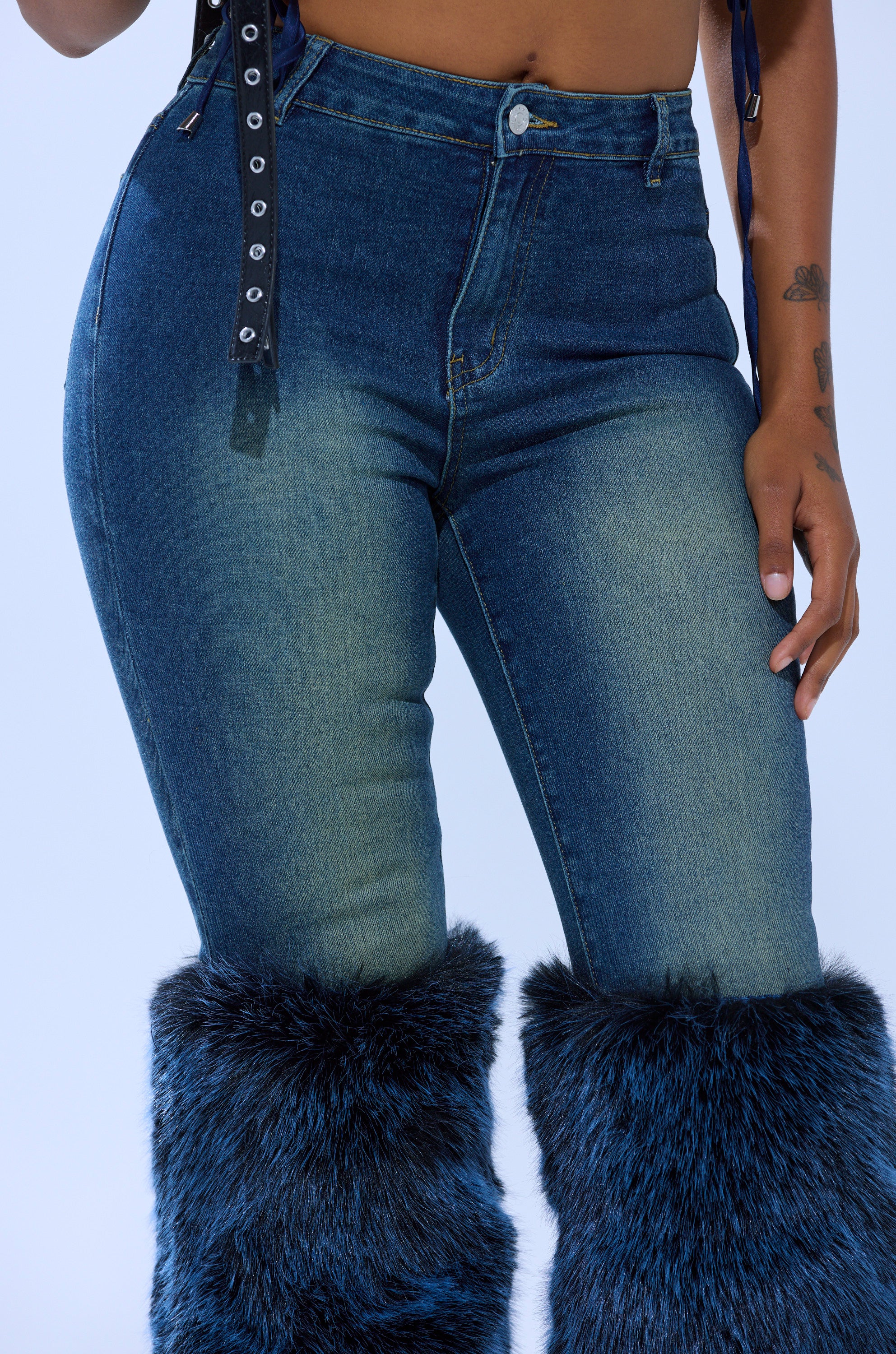 Wearing the ALL A DREAM DENIM PANT WITH FUR CUFFS, a black belt with silver eyelets, and showing part of a tattoo on their arm.