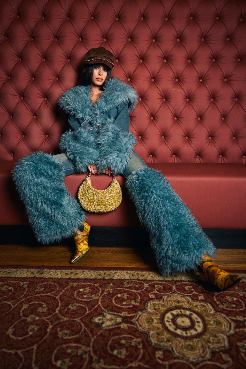 Wearing SIT PRETTY FUR TRIM JEANS IN BLUE DENIM, a person in yellow boots and a brown hat sits on a red tufted bench, holding a circular yellow bag over a patterned red carpet.