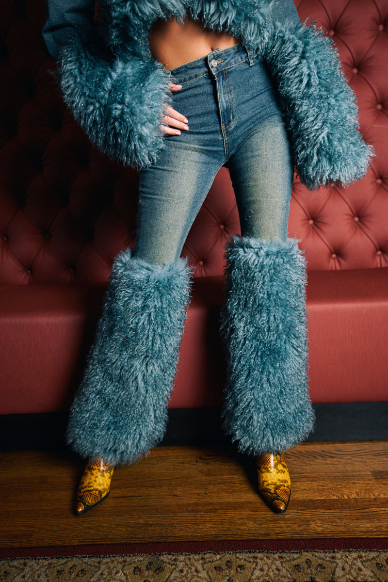 A person models the SIT PRETTY FUR TRIM JEANS IN BLUE DENIM with fluffy blue leg warmers and matching jacket sleeves, standing before a red tufted couch on a wooden floor, highlighting pointed yellow snakeskin boots.