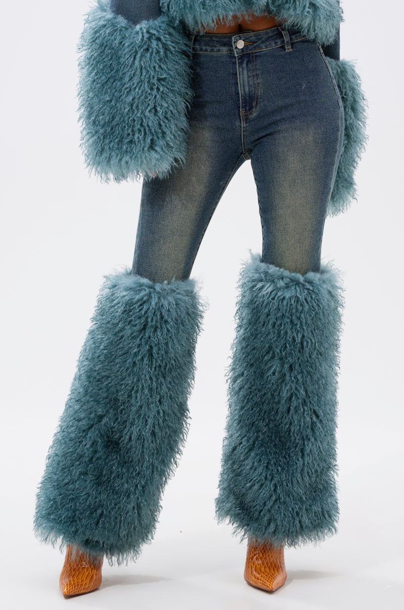 A person wears SIT PRETTY FUR TRIM JEANS IN BLUE DENIM featuring large teal fur trim on the legs and sleeves, paired with orange textured heeled shoes against a plain white background.