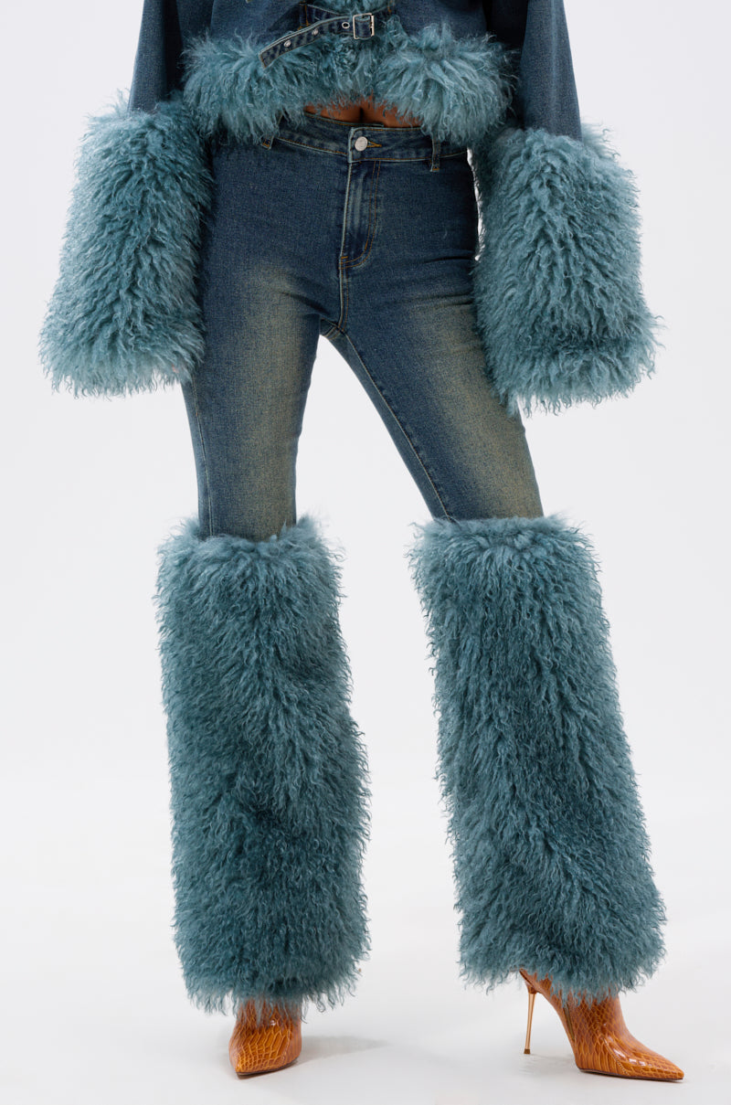 A person models SIT PRETTY FUR TRIM JEANS IN BLUE DENIM, featuring oversized fluffy blue leg warmers and matching sleeve cuffs, paired with brown high heels. The photo is cropped above the shoulders.