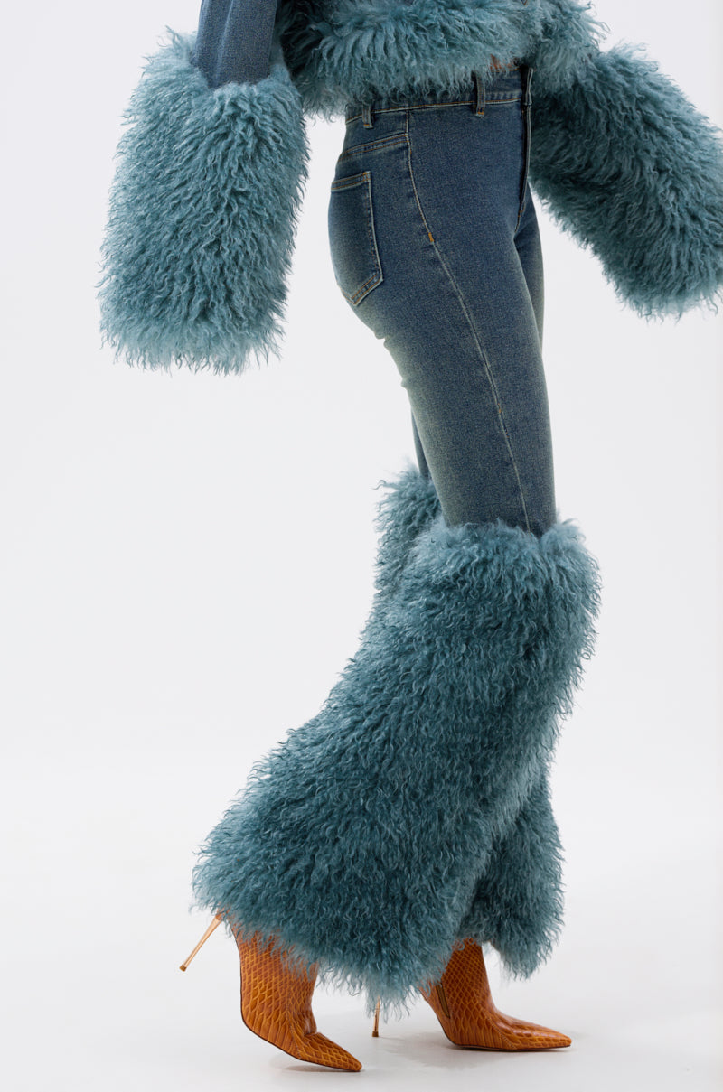 Model wearing SIT PRETTY FUR TRIM JEANS IN BLUE DENIM with shaggy faux fur cuffs, styled with brown high-heeled boots, walking against a plain white background.