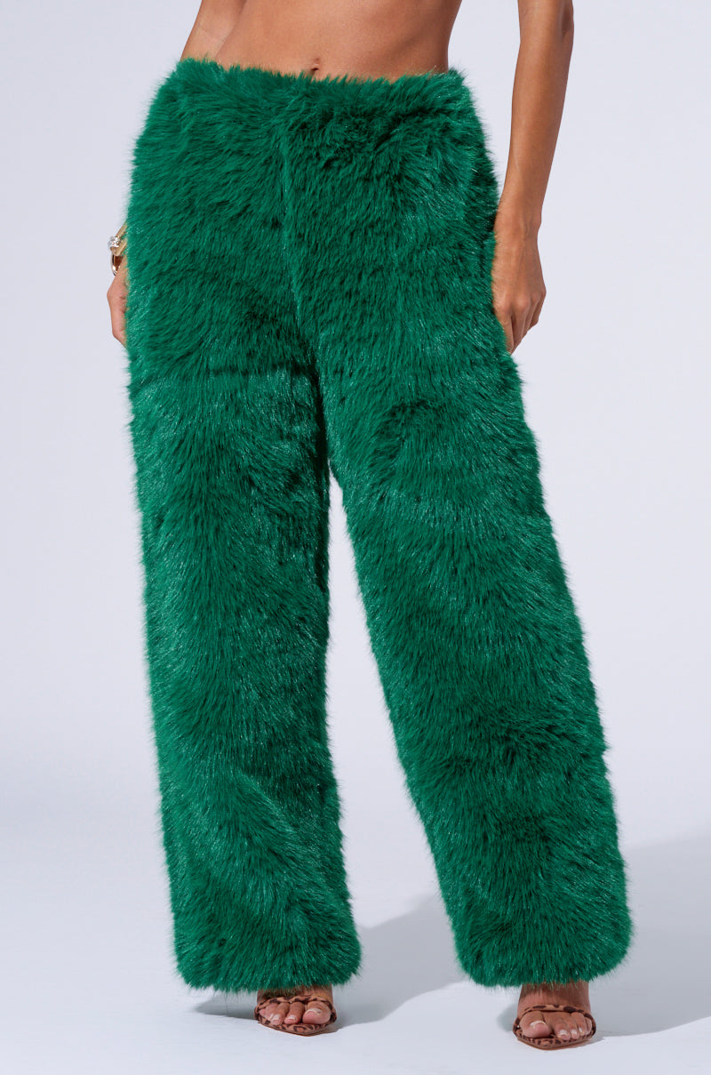 A person is modeling the SEEING GREEN WIDE LEG FASHION FUR PANT, made of vibrant and textured fluffy green faux fur. These high-rise, wide-leg pants stand out against the plain background. The person's midriff and arms are visible, and they are wearing strappy, open-toed sandals.