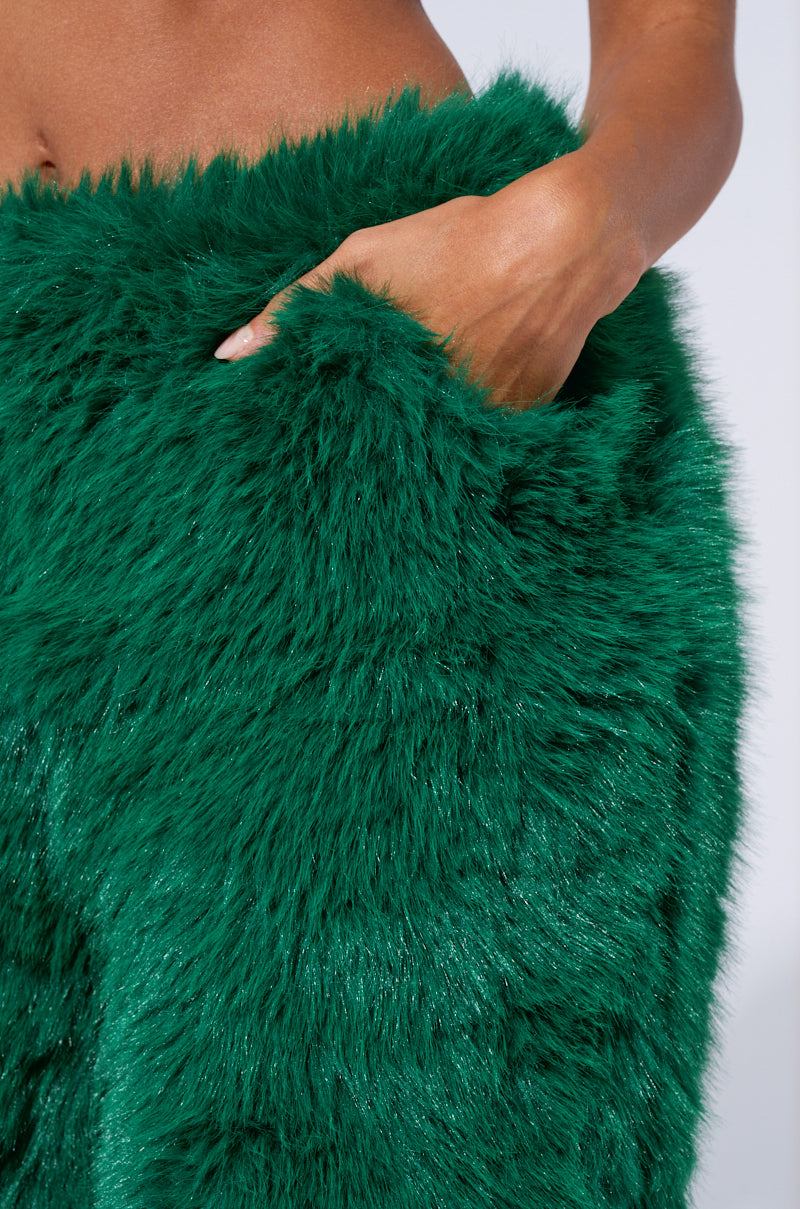 A person with light brown skin showcases the vibrant SEEING GREEN WIDE LEG FASHION FUR PANT, with one hand tucked into a pocket. The texture of the faux fur pants appears soft and plush, with shimmering threads interwoven into the furry material.