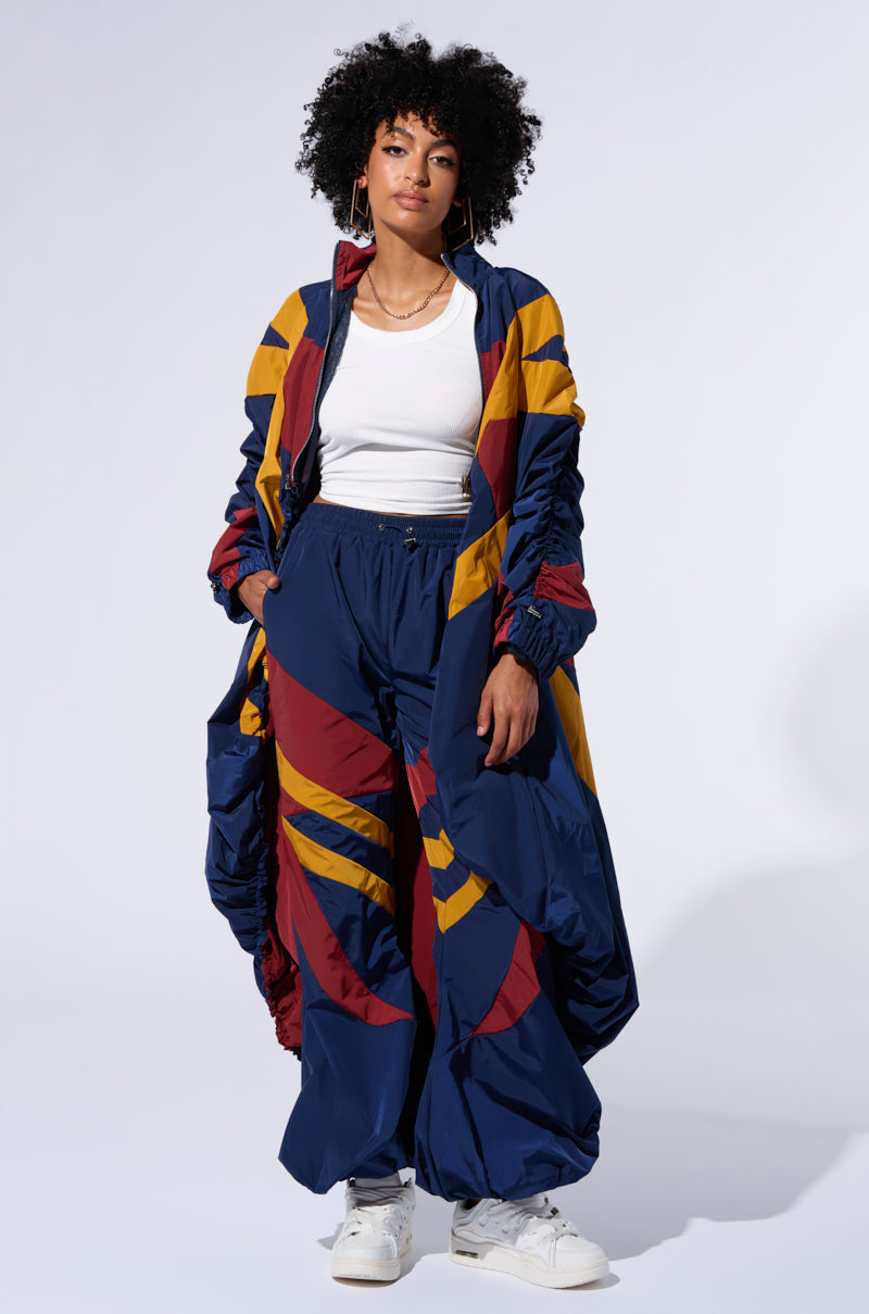 A person with curly hair stands against a plain white background, confidently showcasing the vibrant BORN TO RUN WINDBREAKER TRENCH adorned with blue, maroon, and yellow color block prints. They pair the look with a crisp white tank top, matching white sneakers, and large hoop earrings as they gaze directly at the camera.