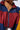 Close-up of a person's torso wearing the BORN TO RUN WINDBREAKER TRENCH, a color-block windbreaker trench with a zipper. The jacket features dark blue, maroon, and yellow sections and an adjustable drawcord. The person is also wearing a thin gold chain necklace and has curly hair. Their face is not visible.