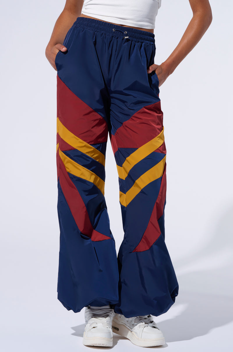 A person wearing the BORN TO RUN WINDBREAKER JOGGERS, featuring navy blue fabric with red and yellow chevron patterns, stands with their hands in their pockets. The person also wears a white sleeveless top and white sneakers. The background is a plain light color.