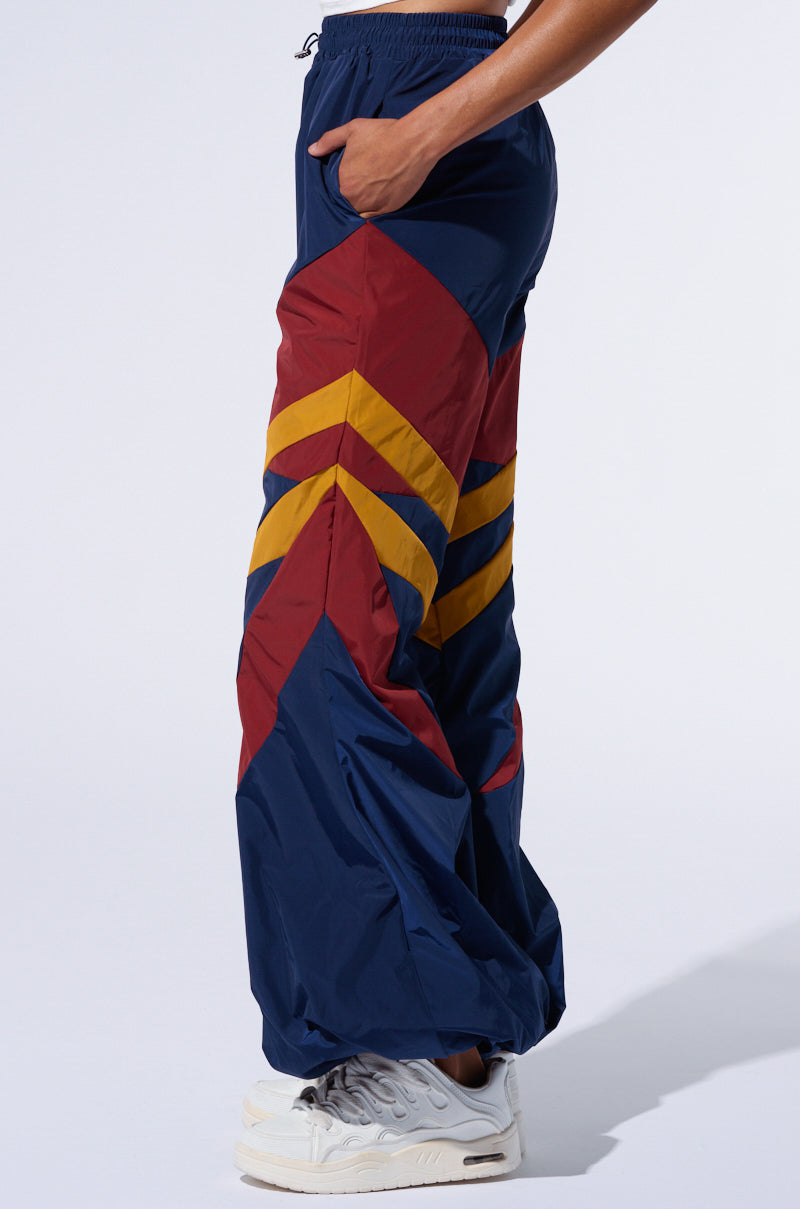 A person is modeling the BORN TO RUN WINDBREAKER JOGGERS, featuring blue athleisure bottoms accented with red and yellow color block paneling along the sides. They are also seen in white sneakers against a white background. The joggers include an elastic waistband and offer a relaxed fit. Only their lower body is shown in the image.