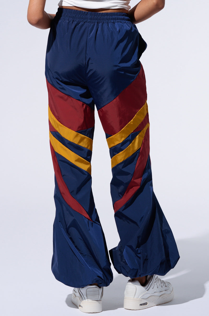 A person is wearing dark blue Born To Run Windbreaker Joggers, which boast red and yellow geometric patterns on the back. These joggers include an elastic waistband and cuffs, color block paneling, and pair perfectly with white sneakers for a stylish athleisure look.