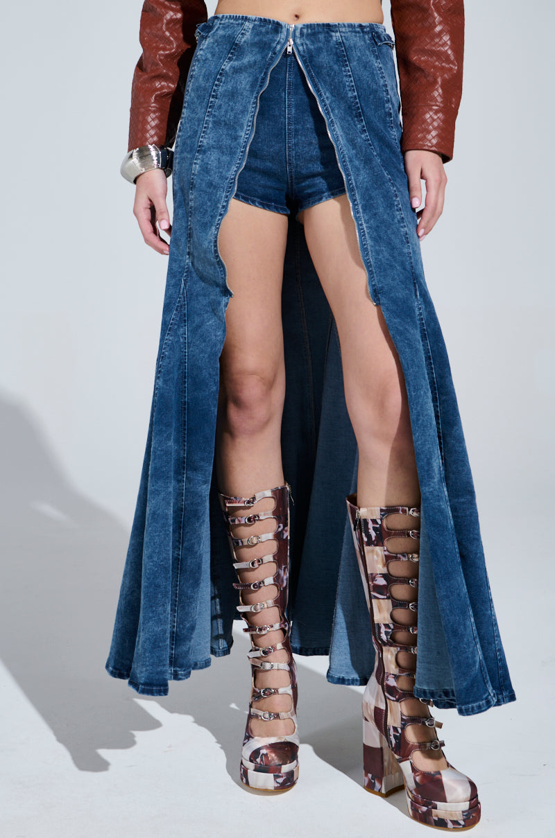 Wearing a Painted Lady Maxi Skirt with built-in shorts, they pair it with a brown leather jacket and brown patterned knee-high platform boots. A large silver cuff bracelet adorns their left arm as they stride confidently.