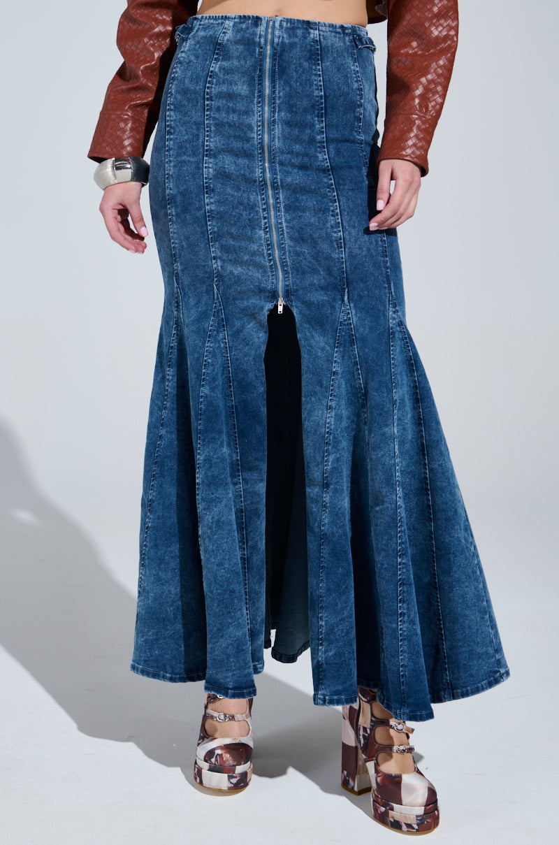 A person is wearing the PAINTED LADY MAXI SKIRT WITH BUILT IN SHORT, a long blue stretchy denim maxi skirt featuring a front zipper, high-rise design with a slit and flare. It's paired with a brown textured leather jacket and platform shoes with mixed patterns.