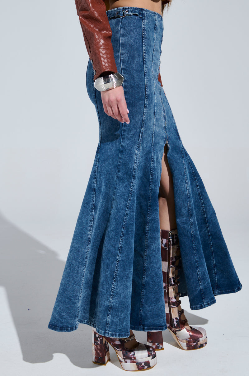 Modeling the Painted Lady Maxi Skirt with built-in shorts, featuring high-waisted stretchy blue denim and a front slit, someone pairs it effortlessly with a brown jacket and patterned platform boots. A chunky silver bracelet adorns one wrist, all set against a plain gray background.
