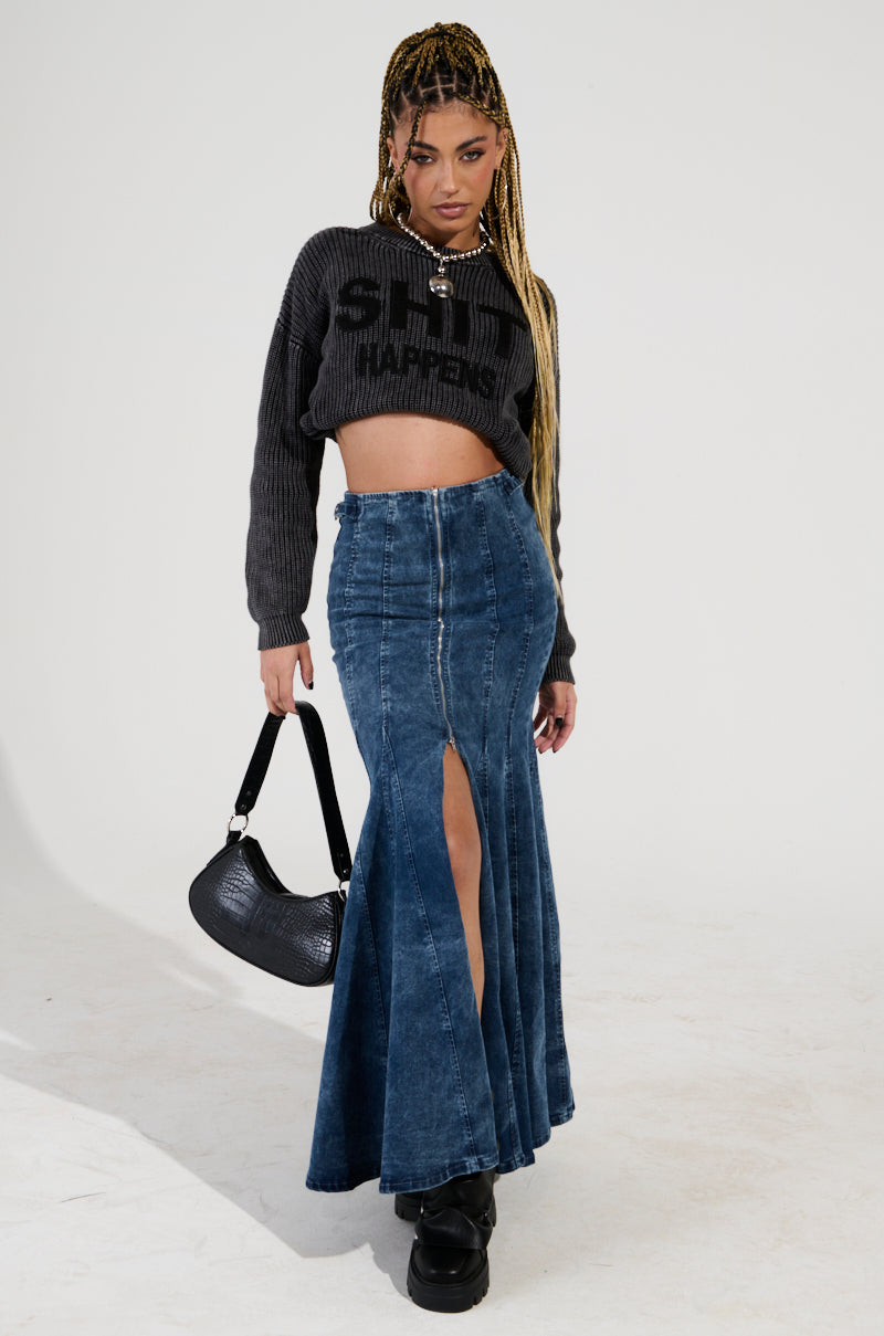 A person with long braided hair stands posed against a white background, wearing a cropped gray sweater and the PAINTED LADY MAXI SKIRT WITH BUILT IN SHORT. Complemented by black platform shoes and holding a black handbag, their outfit blends chic style and comfort perfectly.