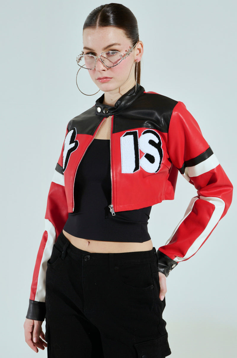 A person wearing the ACE CROPPED GRAPHIC MOTO jacket by AKIRA Label paired with black pants. They have long hair pulled back, large hoop earrings, unique glasses with a chain, and pose confidently with a neutral expression.