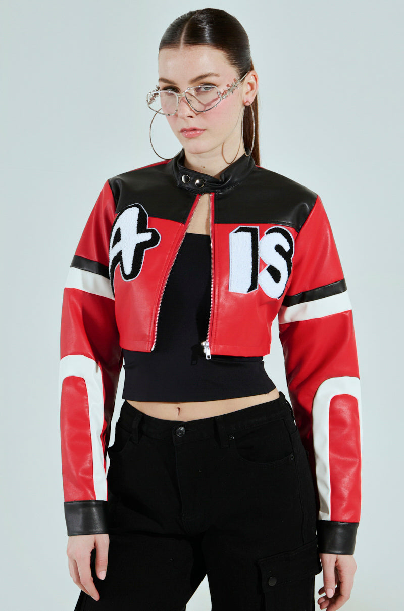 A woman with long hair, oversized glasses, and the ACE CROPPED GRAPHIC MOTO jacket from AKIRA Label pairs it with a black crop top and pants. Standing against a plain background, she exudes confidence as she looks directly at the camera.
