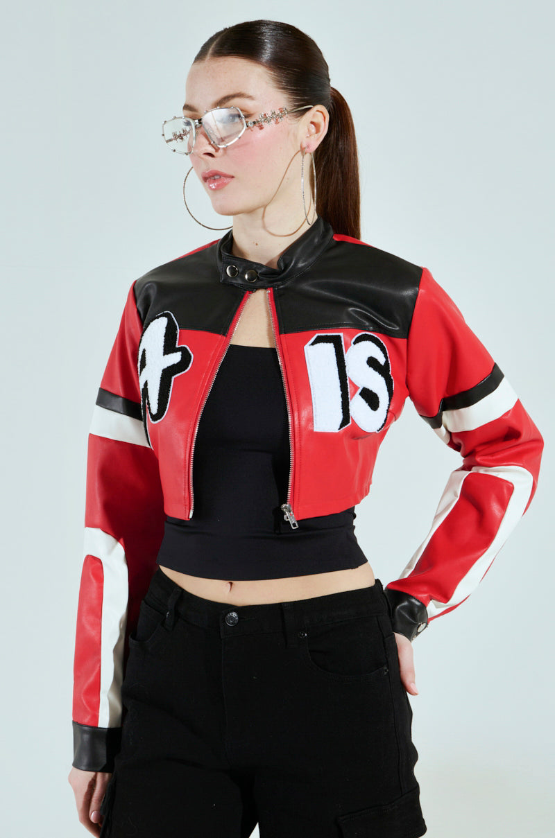 A woman with long dark hair tied back wears oversized sunglasses and large hoop earrings, dressed in a cropped ACE GRAPHIC MOTO jacket with "18" on it, over a black top and high-waisted pants. She stands against a light background.