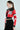 Wearing the ACE CROPPED GRAPHIC MOTO jacket, which features bold red and black faux leather with white lettering, a person pairs it with black pants, large hoop earrings, and futuristic clear glasses. Their hair is styled in a sleek ponytail against a plain light-colored background.