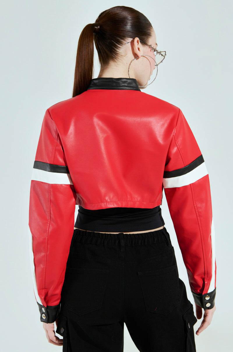 A person with long hair in a ponytail, turned away, is wearing the AKIRA Label ACE Cropped Graphic Moto Jacket in red faux leather with black and white striped sleeves and a black collar, paired with black pants and large hoop earrings.