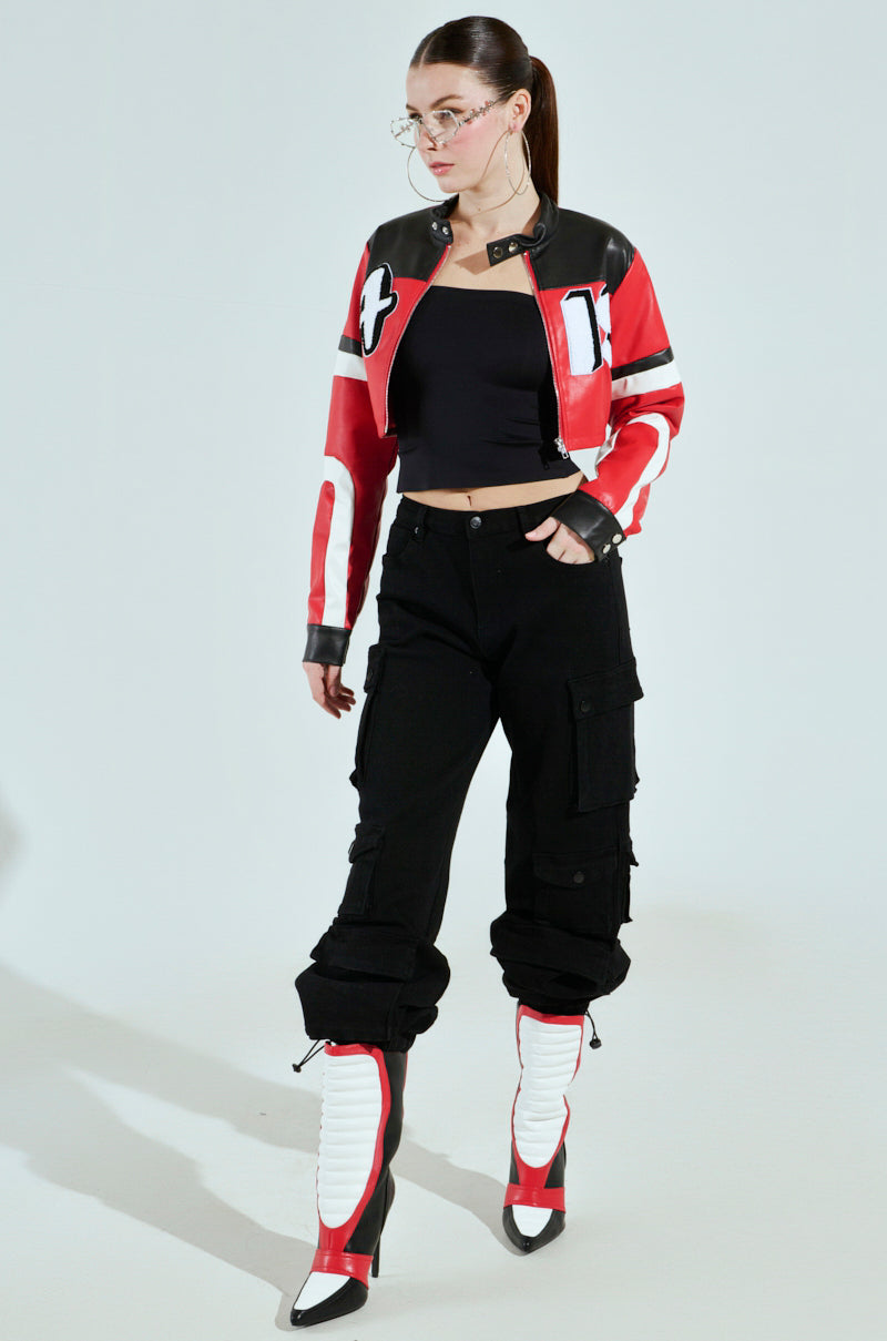 A person confidently stands in an AKIRA Label ACE CROPPED GRAPHIC MOTO jacket, black top, and cargo pants. They wear high-heeled boots with white accents and large glasses against a light-colored background.
