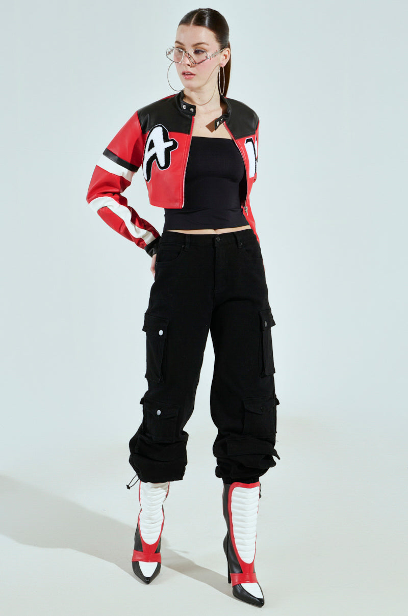 Wearing the ACE CROPPED GRAPHIC MOTO red and black varsity-style jacket, a person pairs it with a black top, cargo pants, and white knee-high boots with black pointed toes. They accessorize with large hoop earrings and glasses, striking a confident side pose with hands on hips.