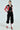 Wearing the ACE CROPPED GRAPHIC MOTO red and black varsity-style jacket, a person pairs it with a black top, cargo pants, and white knee-high boots with black pointed toes. They accessorize with large hoop earrings and glasses, striking a confident side pose with hands on hips.