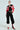 Dressed in the ACE CROPPED GRAPHIC MOTO jacket in red and black with white accents, this look includes black cargo pants and high-heeled boots with red and white stripes, accessorized with hoop earrings and sunglasses, all set against a plain white background.