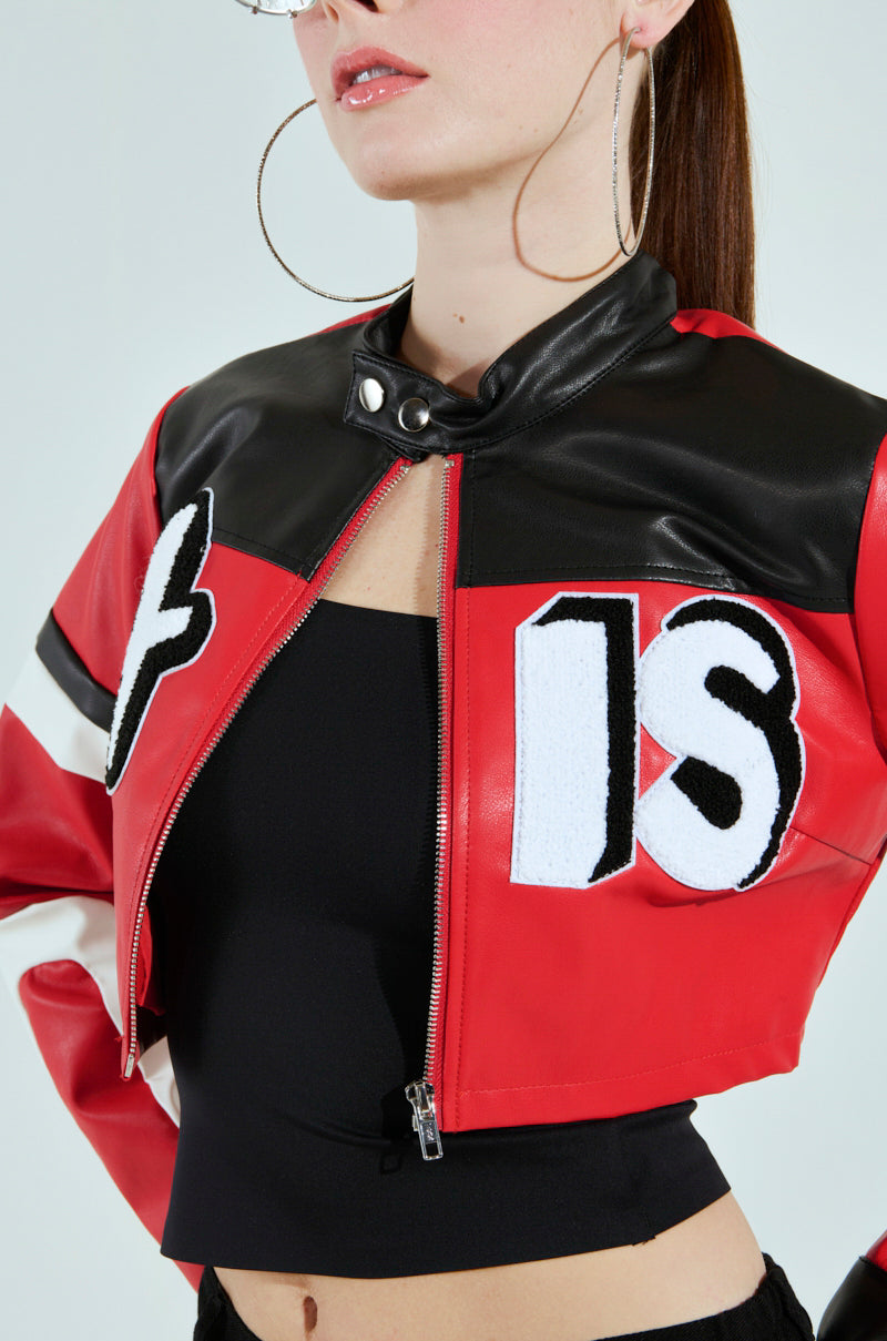 A person in a red and black ACE CROPPED GRAPHIC MOTO jacket with large white and black patches, hoop earrings, and a black top. The AKIRA Label jacket features "IS" and a plus sign on the front.