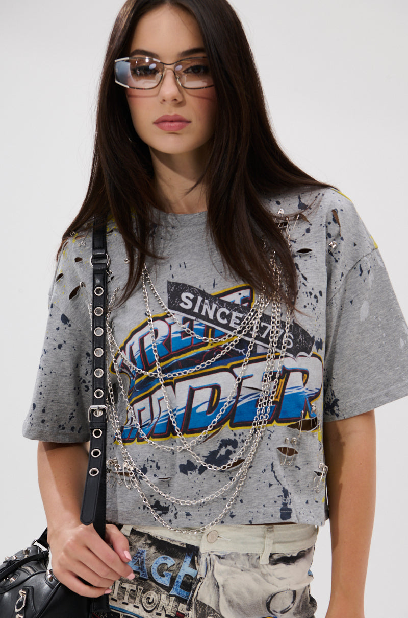 A woman with long dark hair and glasses wears the EXTRA OVERSIZED TEE with layered silver chains and holds a black bag, creating an edgy, punk-inspired look against a plain light-colored background.