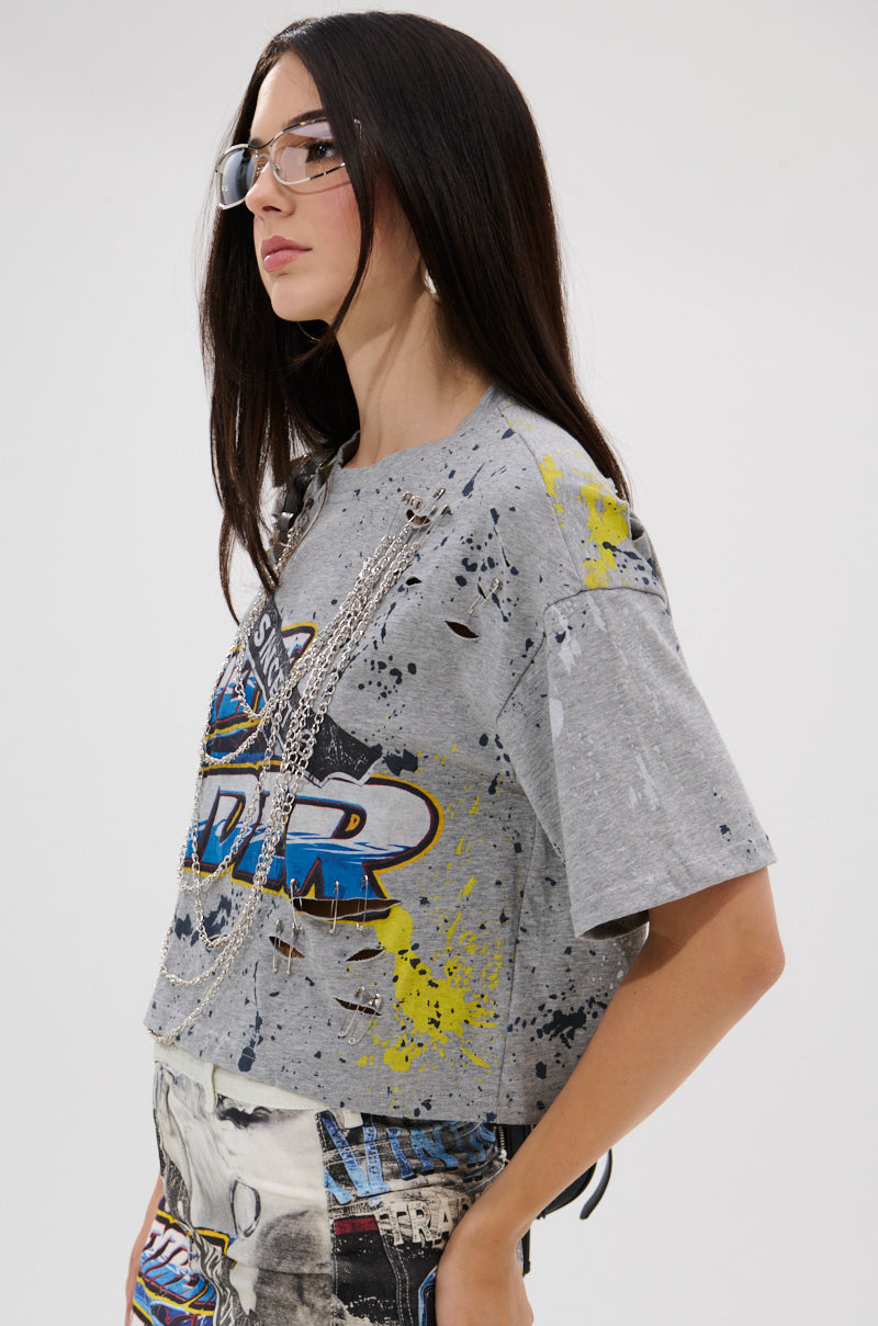 A woman with long dark hair, clear glasses, and layered silver necklaces stands in profile against a plain background, wearing the EXTRA OVERSIZED TEE—a gray distressed t-shirt with blue and yellow accents.