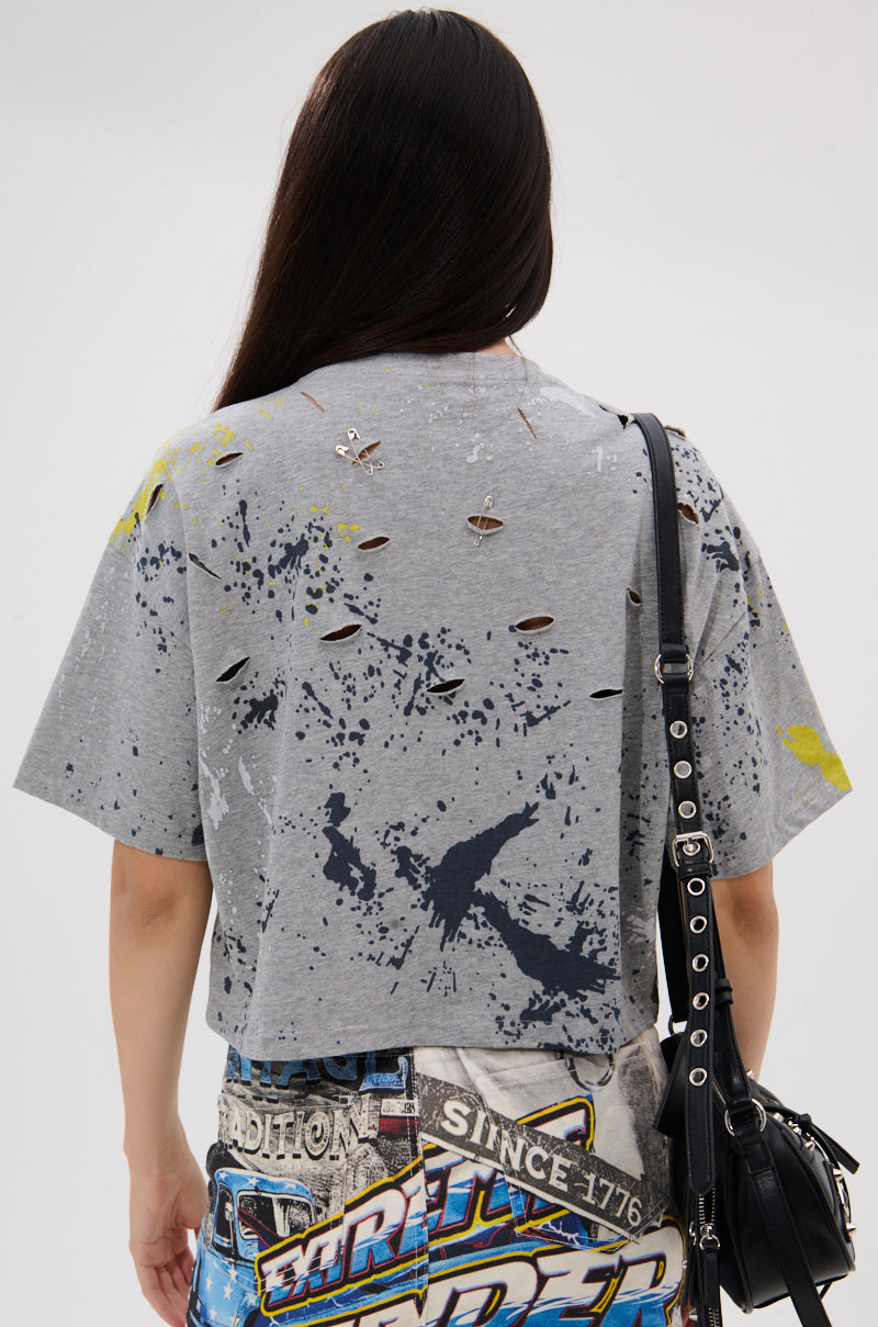 Seen from behind, a person with long dark hair wears the EXTRA OVERSIZED TEE featuring holes and splatter designs, paired with printed blue jeans and a shoulder bag with metal chain details.