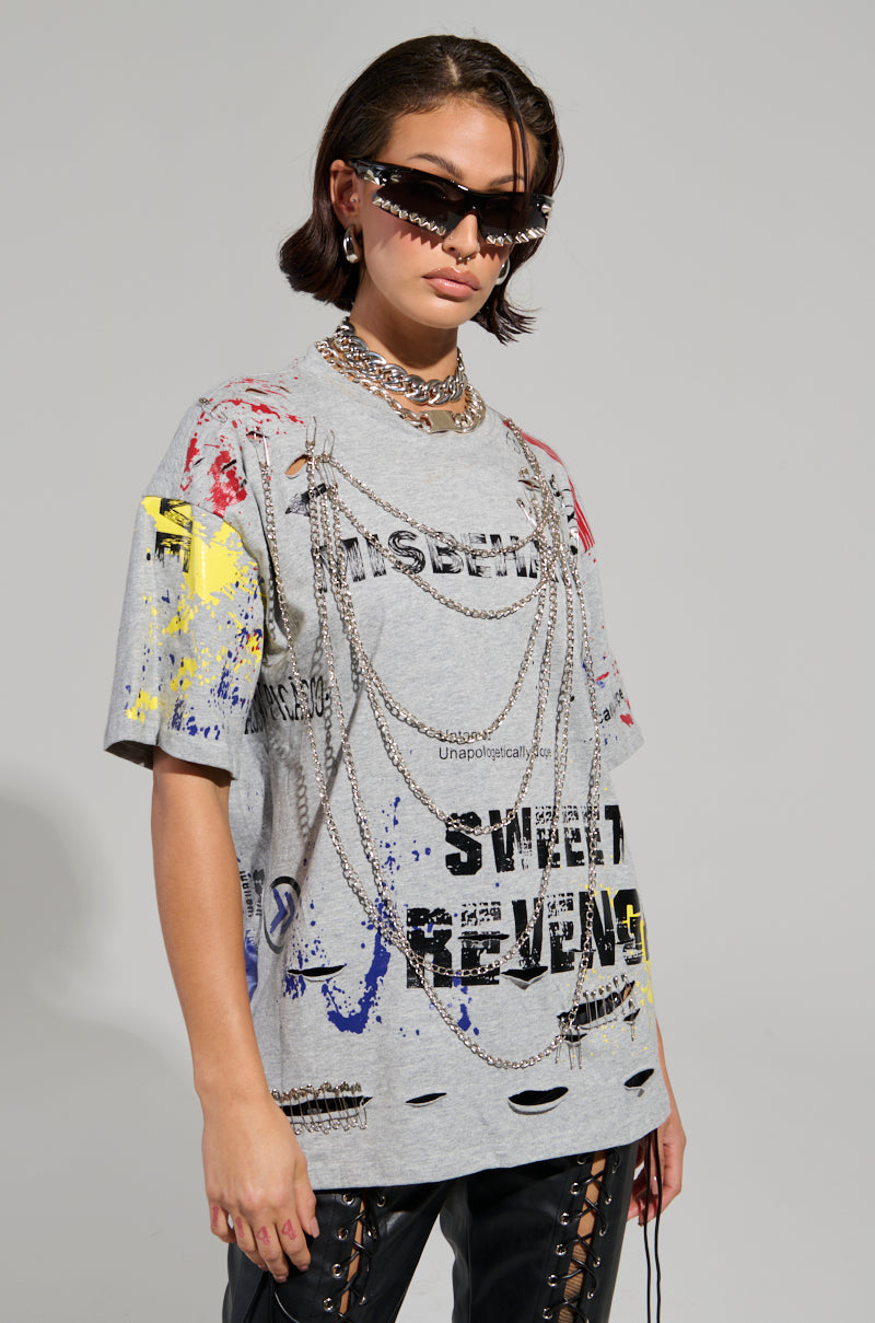 A person sports the SWEET REVENGE DISTRESSED T-SHIRT IN GREY, featuring colorful paint splatters and text on a cotton knit base with ripped details. They enhance the grunge-chic look with layered chain necklaces, sunglasses, and their short, dark hair.