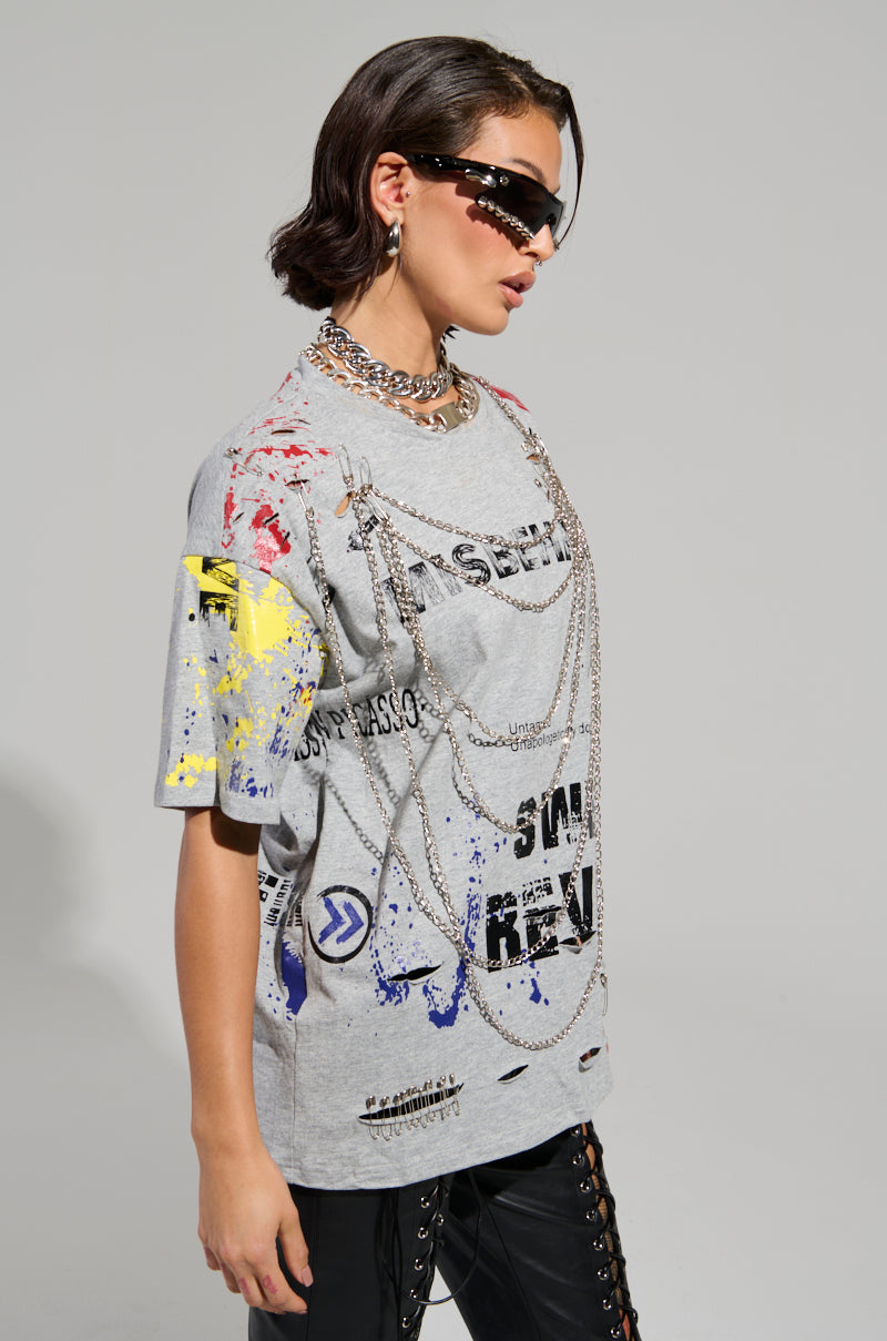 A person clad in the SWEET REVENGE DISTRESSED T-SHIRT IN GREY, featuring vibrant patterns in a gray grunge-chic style, paired with black sunglasses, chains, and black pants embellished with side lacing, embodies a bold and edgy vibe against a plain light gray background.