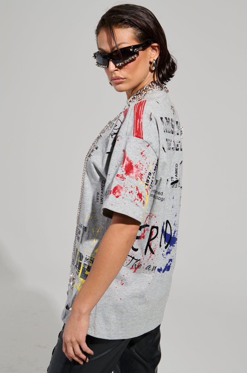 A person dons the SWEET REVENGE DISTRESSED T-SHIRT IN GREY, featuring colorful abstract designs, paired with black pants. With slicked-back hair, dark sunglasses, and a chain necklace, they exude an edgy, modern fashion vibe against a plain backdrop.