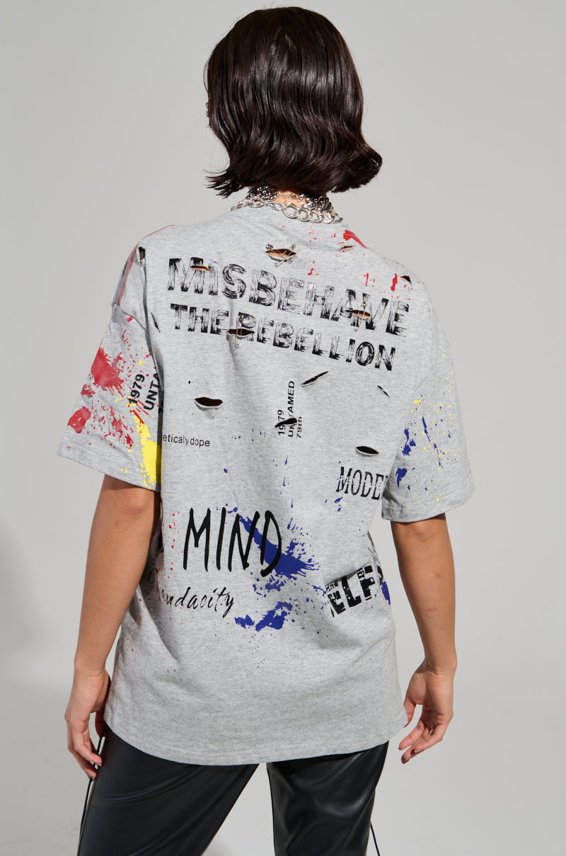 A person wearing the SWEET REVENGE DISTRESSED T-SHIRT IN GREY showcases abstract paint splatters and phrases such as "MISBEHAVE" and "THE REBELLION" on the back. The cotton knit top pairs perfectly with their black pants, as they face away with shoulder-length dark hair.