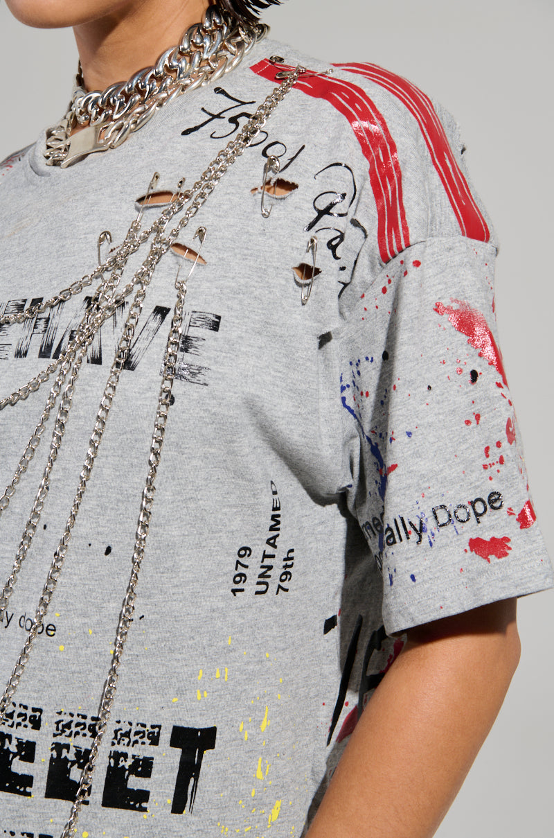 A person is wearing the SWEET REVENGE DISTRESSED T-SHIRT IN GREY, featuring red and black designs with text. This distressed cotton knit graphic T-shirt includes cut-out details, layered chains, and phrases like "UNTA" and "1979 UNTA," exuding an edgy, grunge-chic style.