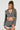 A person with long braided hair stands against a plain background, wearing the Short n Sweet Cropped Blazer, paired with matching high-waisted shorts featuring ruffled edges. They accessorize with large pearl earrings and a necklace, while holding a textured clutch.