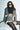 A woman in sunglasses poses against a white brick wall, wearing the SHORT N SWEET CROPPED BLAZER in gray plaid with ruffle details, a matching skirt, a black turtleneck crop top, and dark tights. Her long hair is styled in loose waves.
