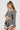 A person with long braided hair wears the SHORT N SWEET CROPPED BLAZER, a stylish gray herringbone wool blend jacket, paired with a matching pleated mini skirt featuring white trim. They accessorize with a large pearl necklace and hoop earrings, set against a plain, light-colored background.