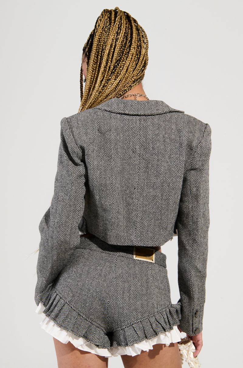 A person with braided hair wearing the SHORT N SWEET CROPPED BLAZER in a gray wool blend herringbone fabric, paired with matching high-waisted shorts adorned with ruffle trim. The cropped blazer design highlights the chic back of the outfit against a plain background.