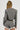 A person with braided hair wearing the SHORT N SWEET CROPPED BLAZER in a gray wool blend herringbone fabric, paired with matching high-waisted shorts adorned with ruffle trim. The cropped blazer design highlights the chic back of the outfit against a plain background.