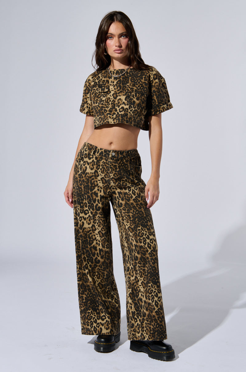 A woman with long brown hair stands against a neutral background, showcasing the stylish CHEETAH SISTERS PANT paired with a matching leopard print cropped T-shirt. The high-waisted pants feature a wide leg silhouette that perfectly complements her black platform shoes, creating a chic and bold look.