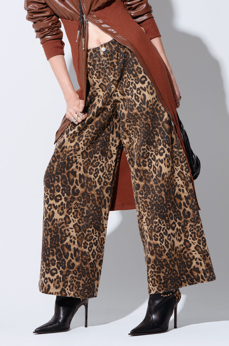 A person is wearing a long brown cardigan with a front zipper, paired with the high-rise, wide-legged CHEETAH SISTERS PANT. They are also wearing black high-heeled ankle boots. The person's hands are adorned with rings and they are carrying a black shoulder bag.