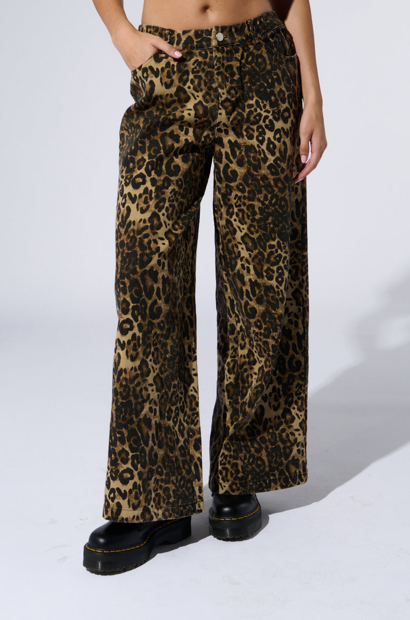 A woman is wearing the CHEETAH SISTERS PANT with a high-rise design and black platform shoes. The pants feature a wide-leg silhouette, giving them a fierce yet stylish look. Her right hand is in her pocket while her left hand rests at her side against the plain, light-colored background.