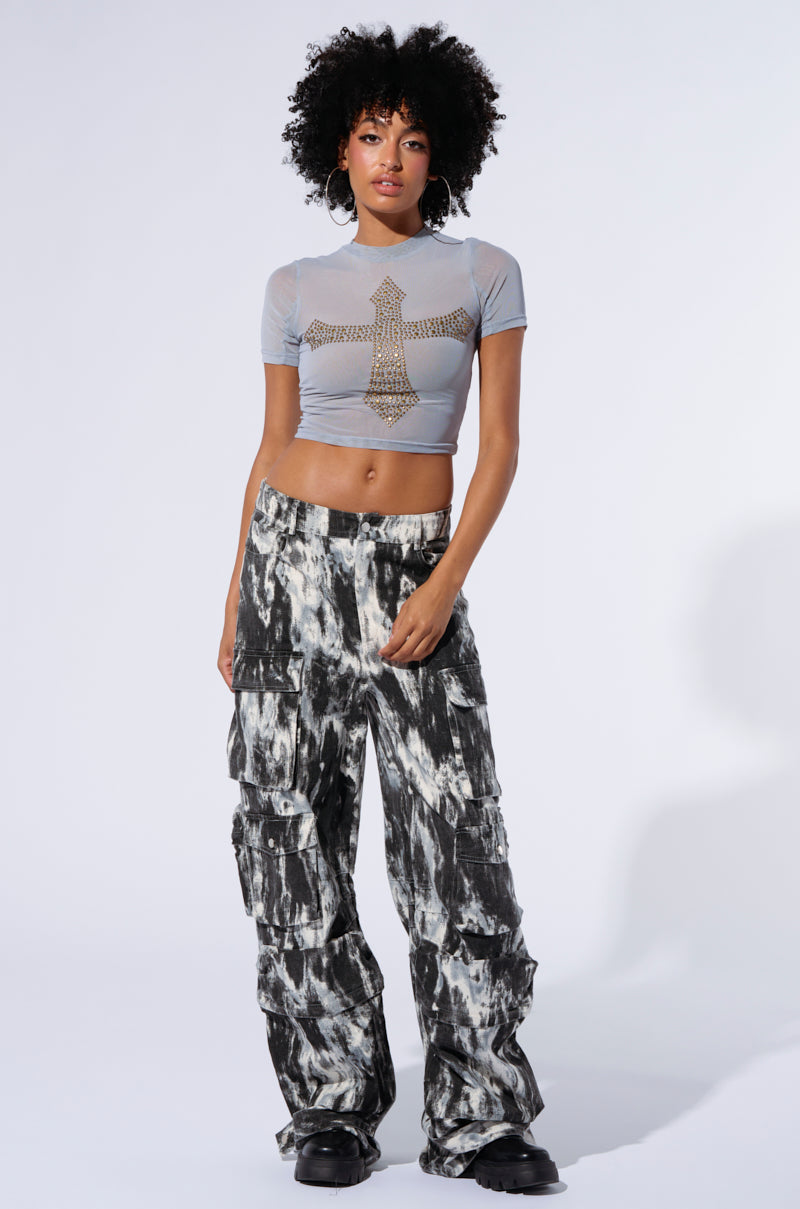 A person with curly hair stands against a white background, wearing a light grey crop top with a bejeweled cross design and loose-fitting, high-rise waist GET WITH IT CARGO PANTS featuring an abstract painterly pattern. They pair the outfit with black boots and accessorize with large hoop earrings.