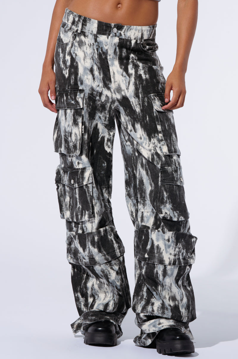 A person is shown wearing the GET WITH IT CARGO PANT, which features a black and white tie-dye design with a high rise waist and multiple pockets. These loose-fitting pants showcase an abstract painterly patterning and are paired with chunky black shoes. The photo is cropped at the waist, focusing on the pants and shoes.