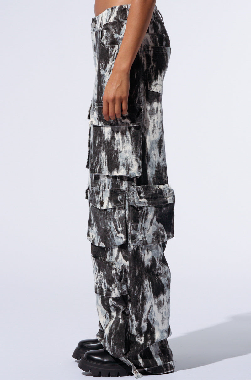 Side profile of a person wearing the GET WITH IT CARGO PANT, which showcases a black and white tie-dye pattern with multiple pockets. These cargo pants offer a loose fit and high-rise waist, paired with chunky black shoes. The background is plain white.