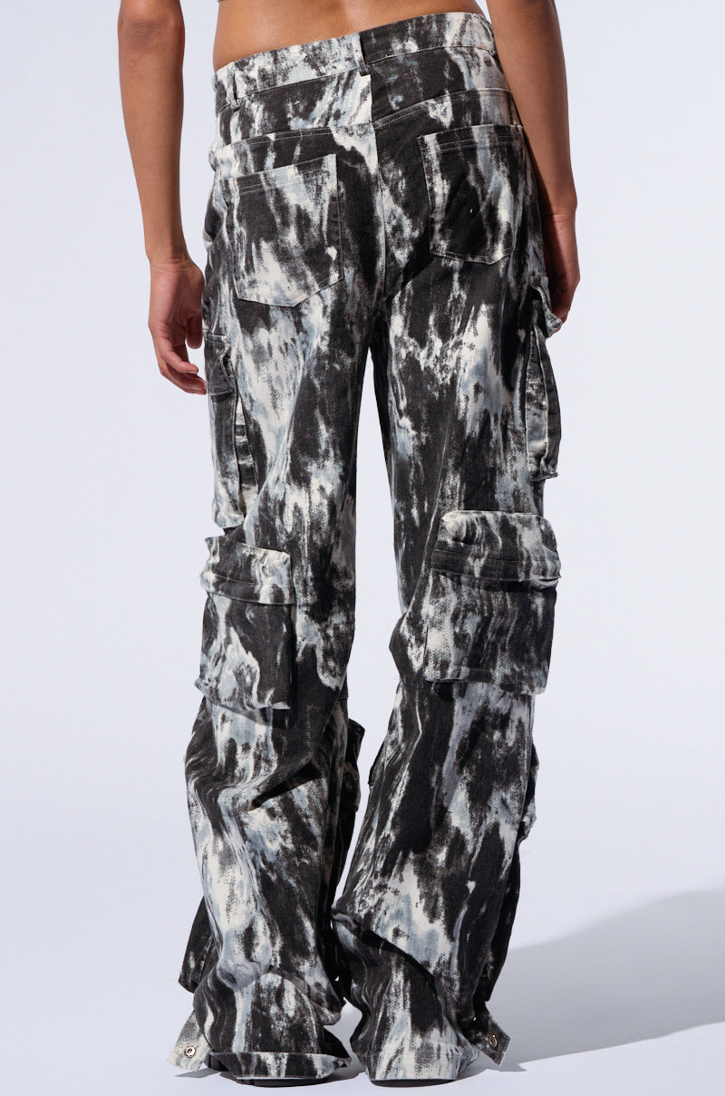 A person is standing with their back to the camera, wearing the GET WITH IT CARGO PANT in black and white tie-dye. The high-rise waist and multiple pockets enhance their practicality, while the abstract painterly patterning provides a striking visual contrast against the plain, light background.
