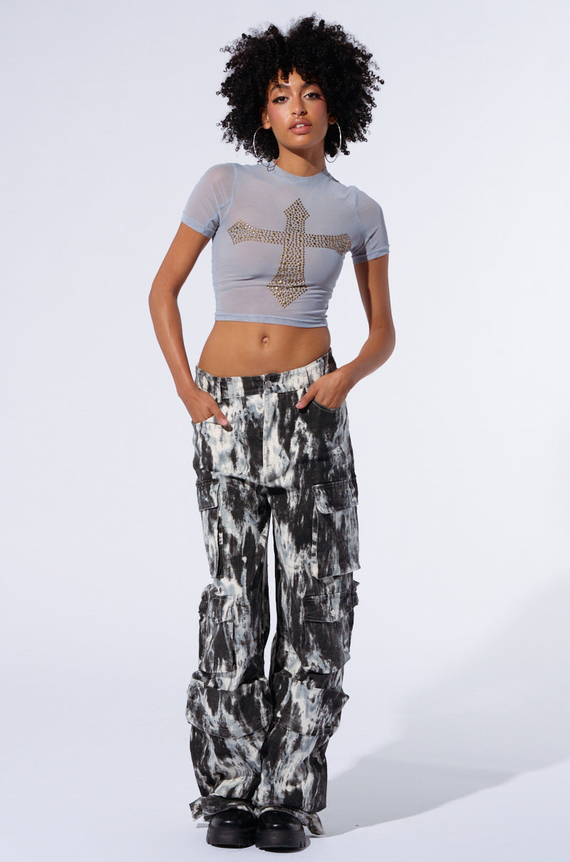A person with curly hair stands against a plain background. They are wearing a light blue crop top with a studded cross design and the GET WITH IT CARGO PANT, which features an abstract painterly pattern in black and white. They have their hands in their pockets and are wearing black shoes.