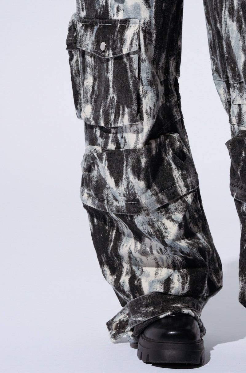 Close-up of a person wearing the GET WITH IT CARGO PANT, showcasing its wide-leg design, high-rise waist, and striking abstract painterly pattern in black and white. The pants flaunt multiple pockets, including one with a secured button flap. The ensemble is completed with black chunky-soled shoes, creating a standout look against a plain light gray background.
